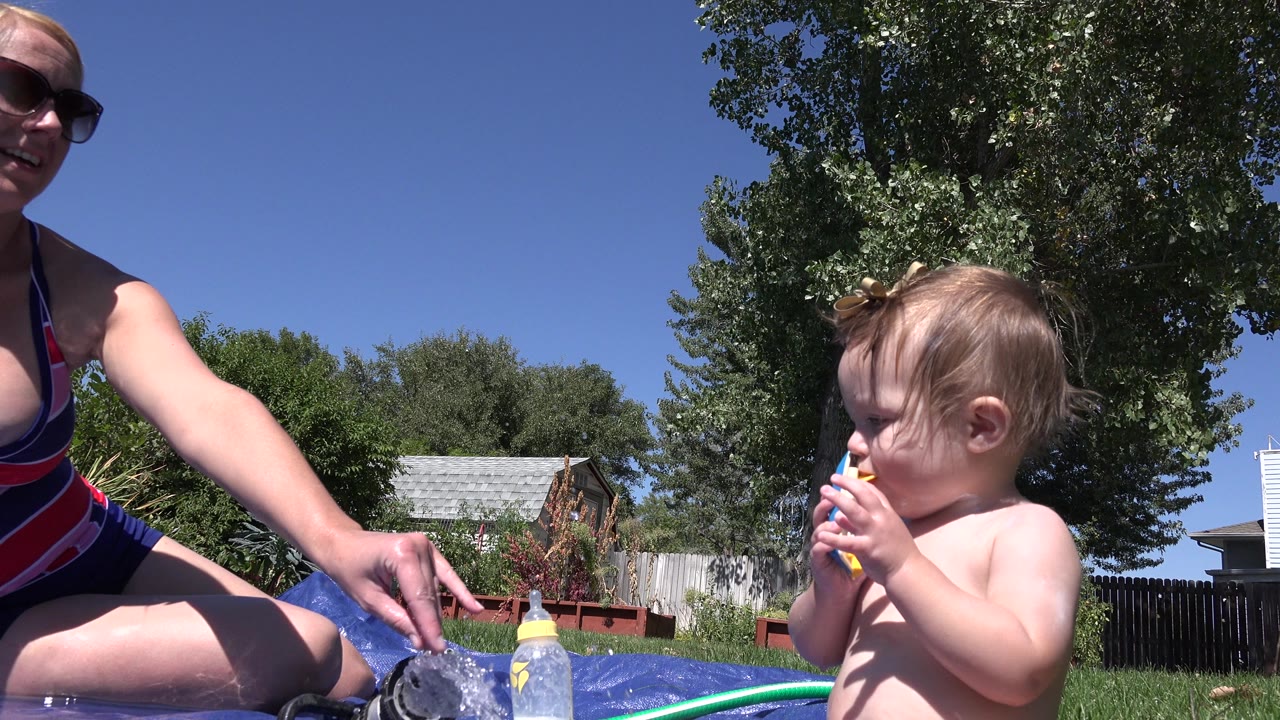 Toddler with toy beside mother on blue splash mat in a sunny backyard.