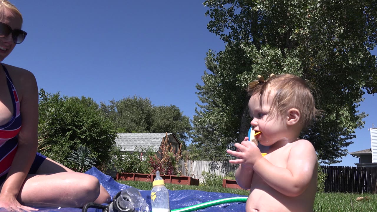 Toddler stands near seated mother with backyard trees behind them.