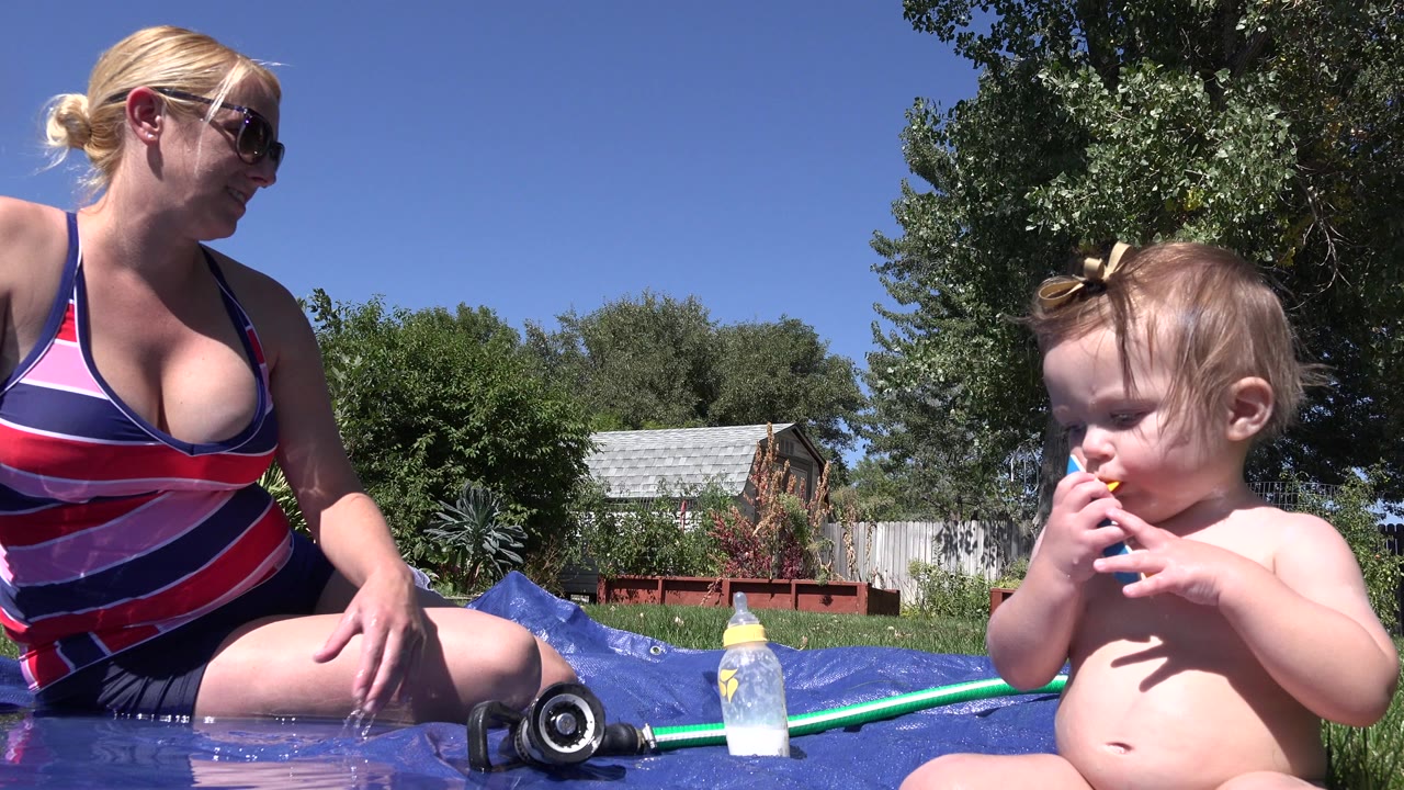 Woman and infant sit on a wet blue surface in a sunny backyard.