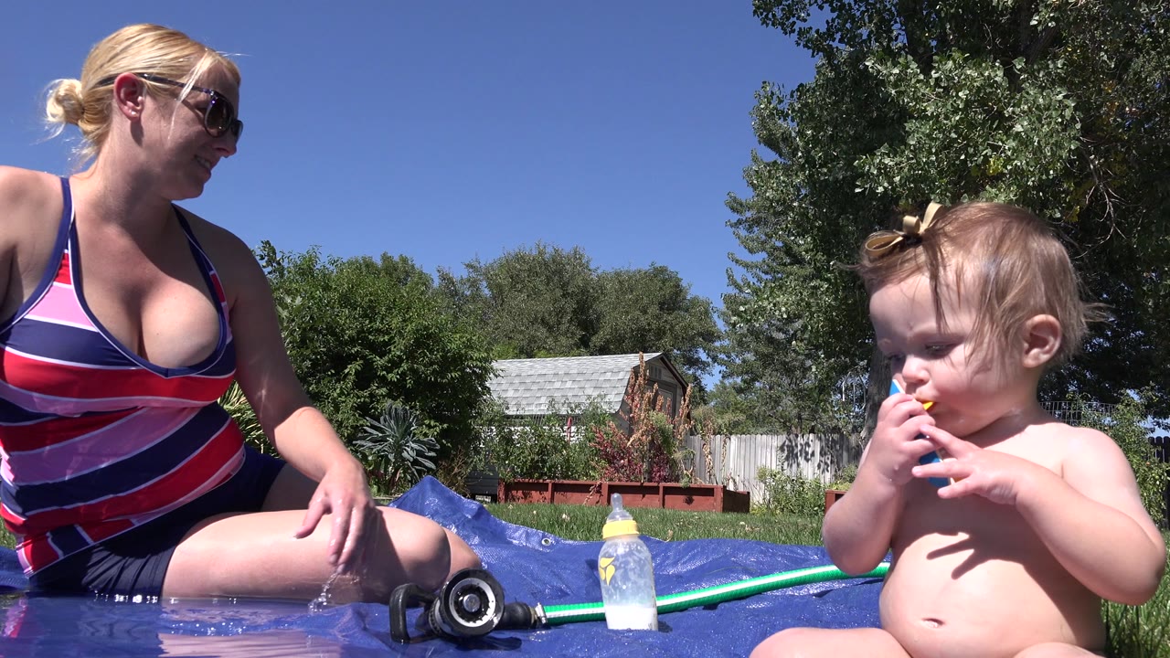The infant keeps a blue and yellow toy near their mouth as the woman sits beside shallow water, with a hose and bottle in the foreground and backyard greenery behind them in Westminster, Colorado.