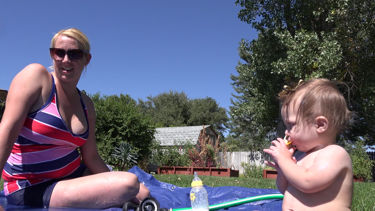 In Westminster, Colorado, a mother in sunglasses and a striped swimsuit sits on a blue tarp while a baby holds a small toy beside grass and backyard trees.