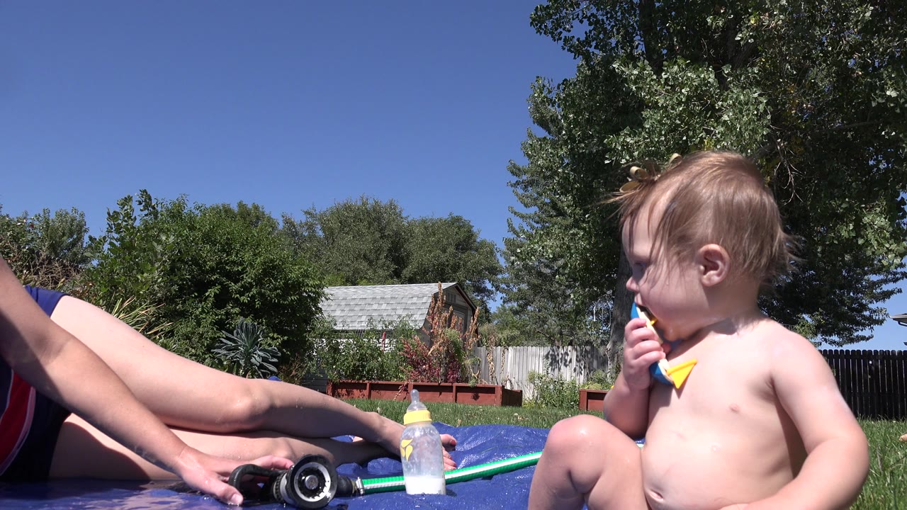 At the start of the Westminster, Colorado clip, a baby sits on a blue mat chewing a toy while an adult's legs, a hose nozzle, a baby bottle, lawn, trees, and a fence are visible.