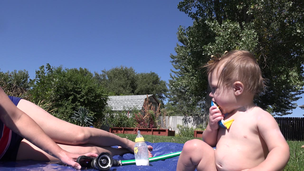 One second into the Westminster, Colorado footage, the baby remains seated at right with the toy near their mouth as the adult's hand stays near the black hose nozzle and bottle on the blue mat.