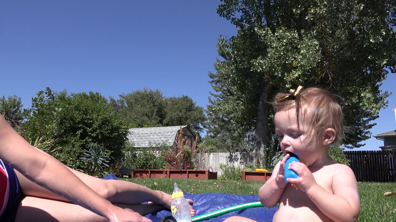 At 0:00 in Westminster, Colorado, a toddler sits on a lawn with a blue water toy near their mouth while a woman's arm and legs, a baby bottle, blue tarp, green hose, trees, fence, and houses are visible.