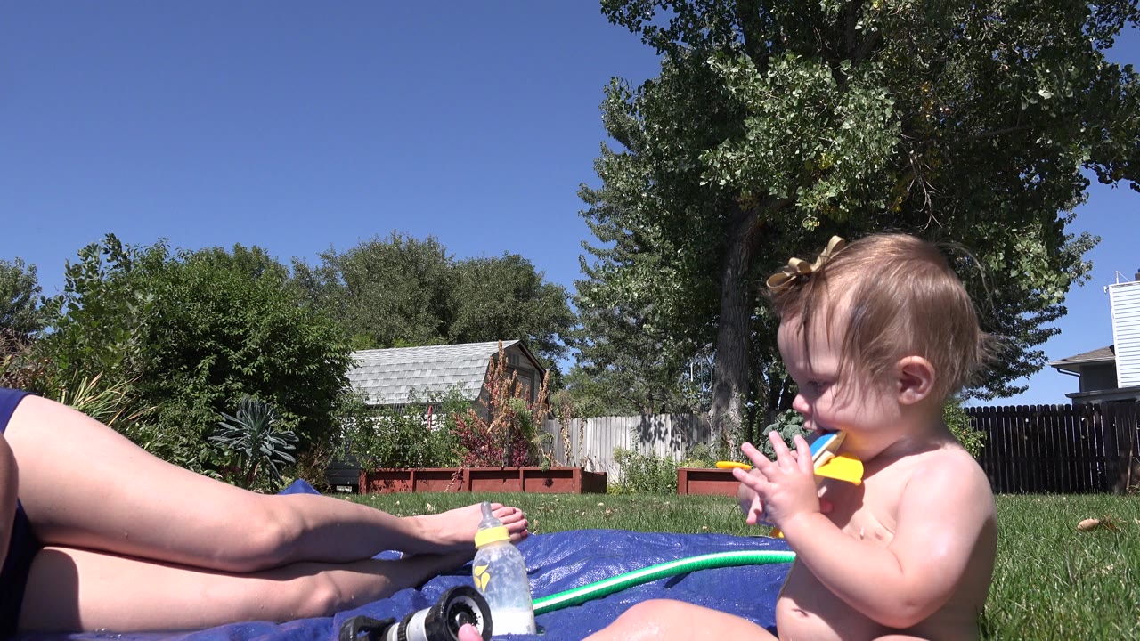 Toddler sitting beside a hose and adult legs in a sunny backyard pool scene.