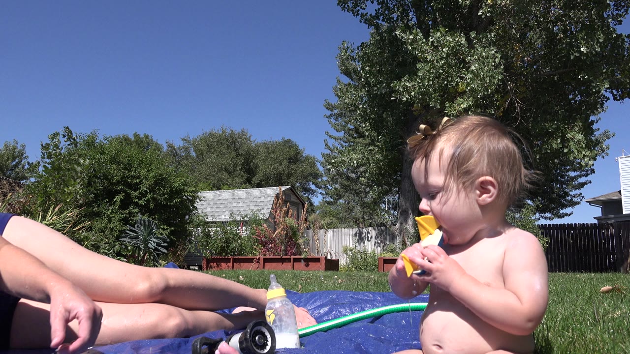 At 0:00 in Westminster, Colorado, a toddler sits on grass beside a blue mat and chews a yellow and blue toy boat while an adult's legs are partly visible at left.