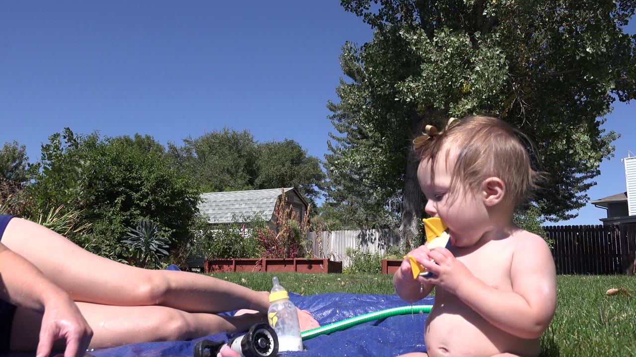 At 0:01 in Westminster, Colorado, the toddler keeps the toy boat at their mouth as a green hose and bottle sit on the blue mat in the sunny lawn foreground.