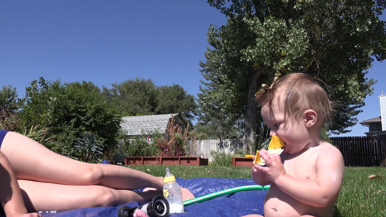 Toddler chewing a toy boat while seated on grass near an adult.