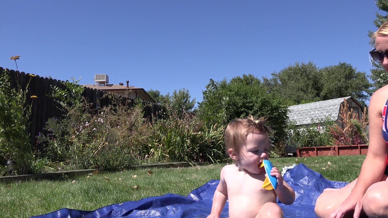 At 0:00 in Westminster, Colorado, a young child sits on blue pool material holding a colorful plastic toy while part of a seated woman in sunglasses appears at the right edge.