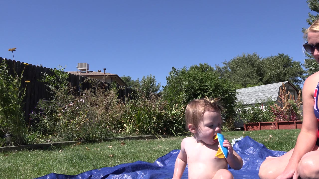 At 0:01 in Westminster, Colorado, the child remains low in frame with a colorful toy near their mouth while the seated woman is cropped at the right edge of the backyard scene.