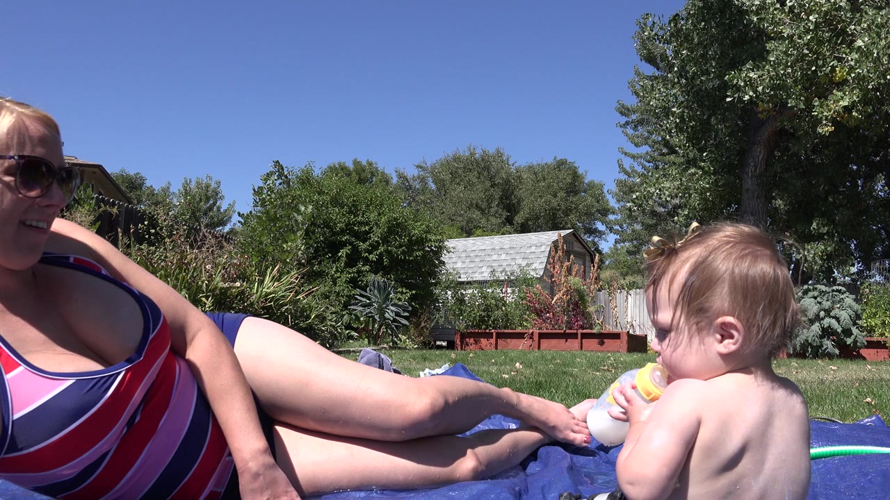 The toddler continues holding the bottle near the woman on the blue mat, with lawn, shrubs, trees, and a small roofed structure visible behind them in Westminster, Colorado.