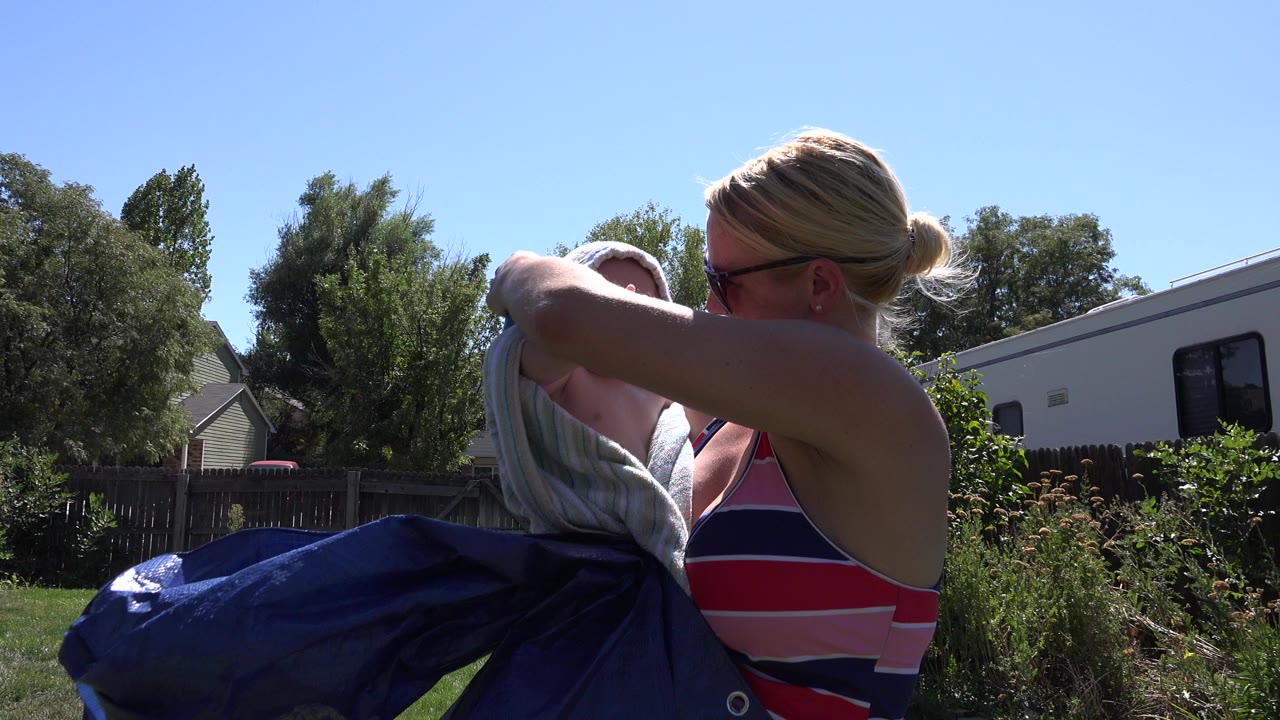 Woman in sunglasses leaning over a baby wrapped in a blue and white towel outdoors.