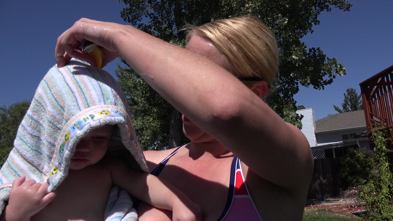 In Westminster, Colorado, a blonde woman in a striped swimsuit lifts a pale hooded towel over a young child beside trees and residential houses.