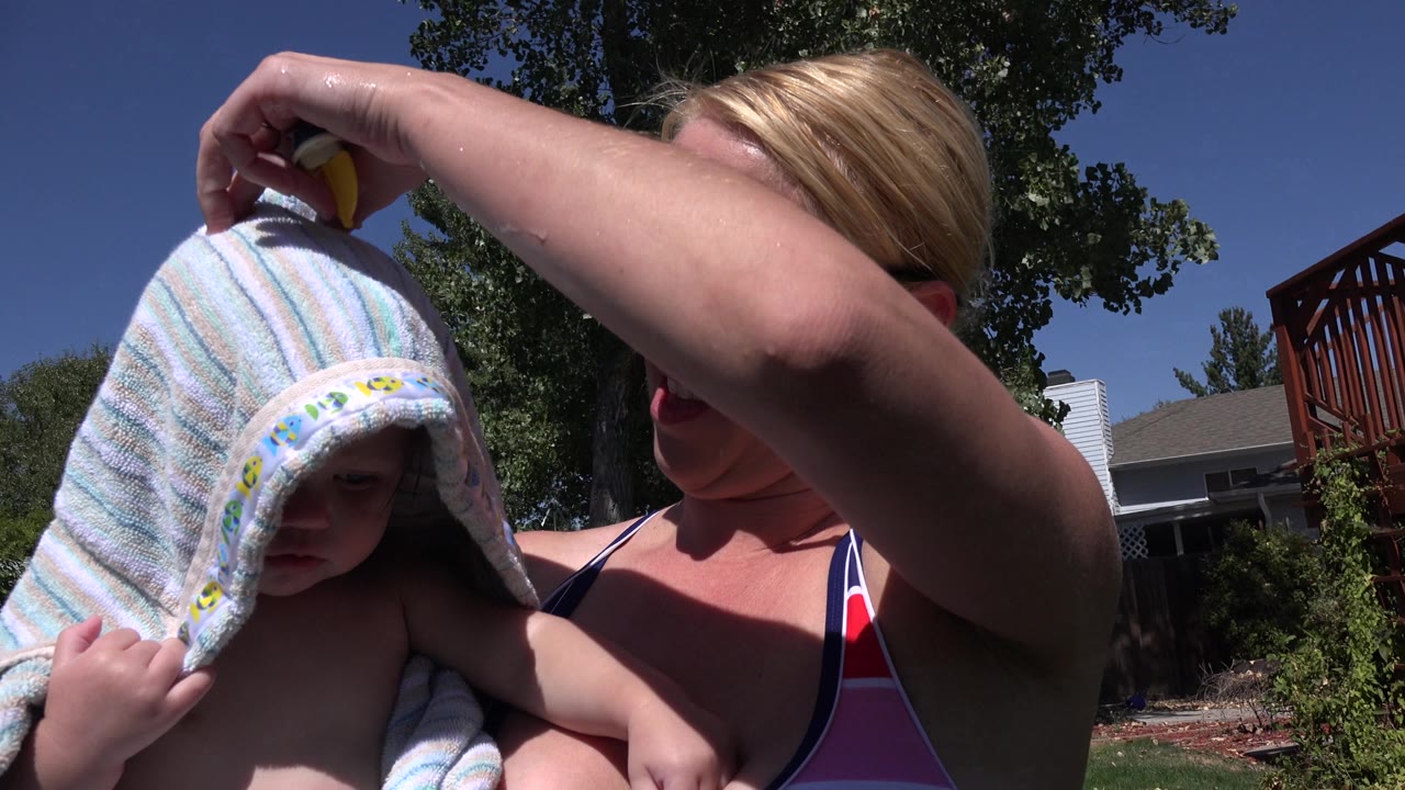 The close view shows the woman partly hidden behind her raised arm while the child looks downward under the striped towel in a Westminster residential yard.