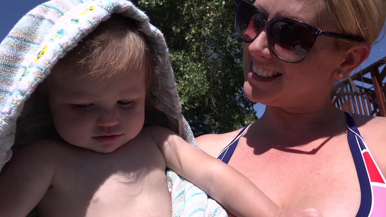 In Westminster, Colorado, a baby with a striped towel over the head sits close to a smiling woman in sunglasses under bright sun, with trees behind them.
