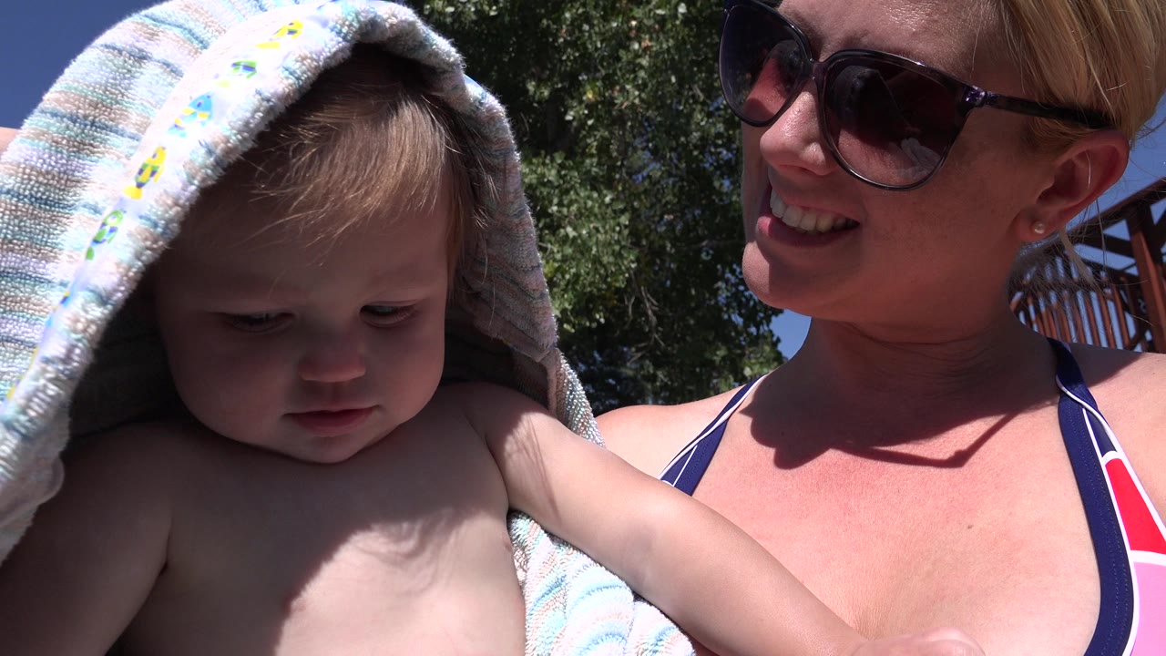In Westminster, Colorado, the baby looks down from under the towel while the woman smiles beside the child, with a dark tree canopy and blue sky in the background.