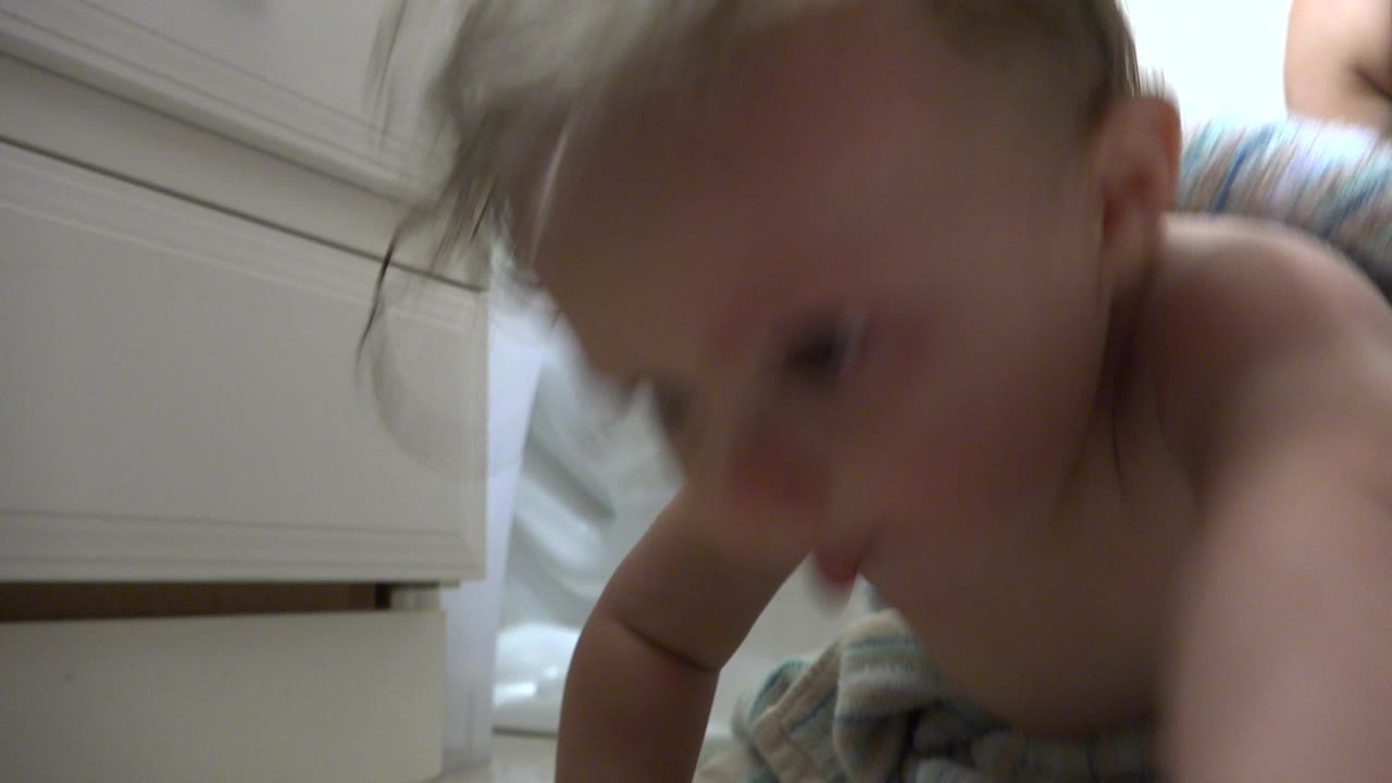 The baby leans forward in soft focus beside white cabinets, with bare shoulders and damp hair visible in the bathroom setting.