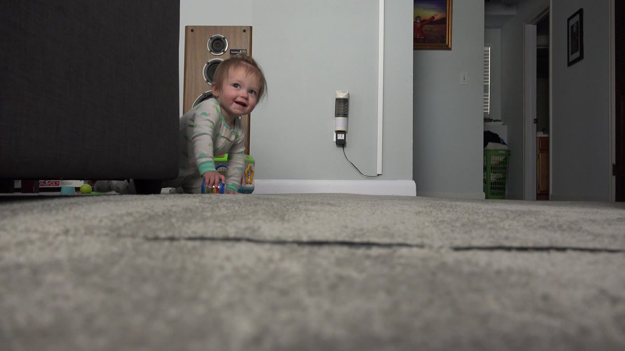 At 0:00 in Westminster, Colorado, a baby in a green and white sleeper crawls partly out from behind a dark couch on a grey carpeted floor.