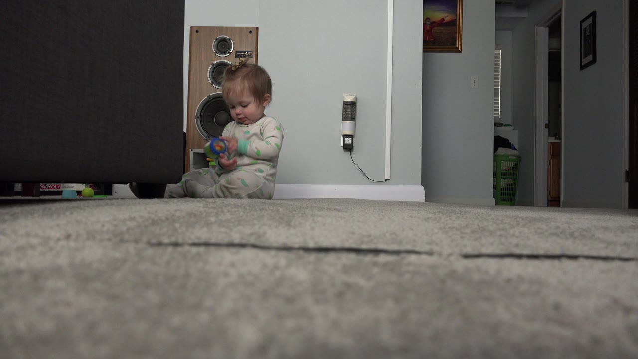 Baby in a green and white sleeper sits on carpet beside a large speaker.