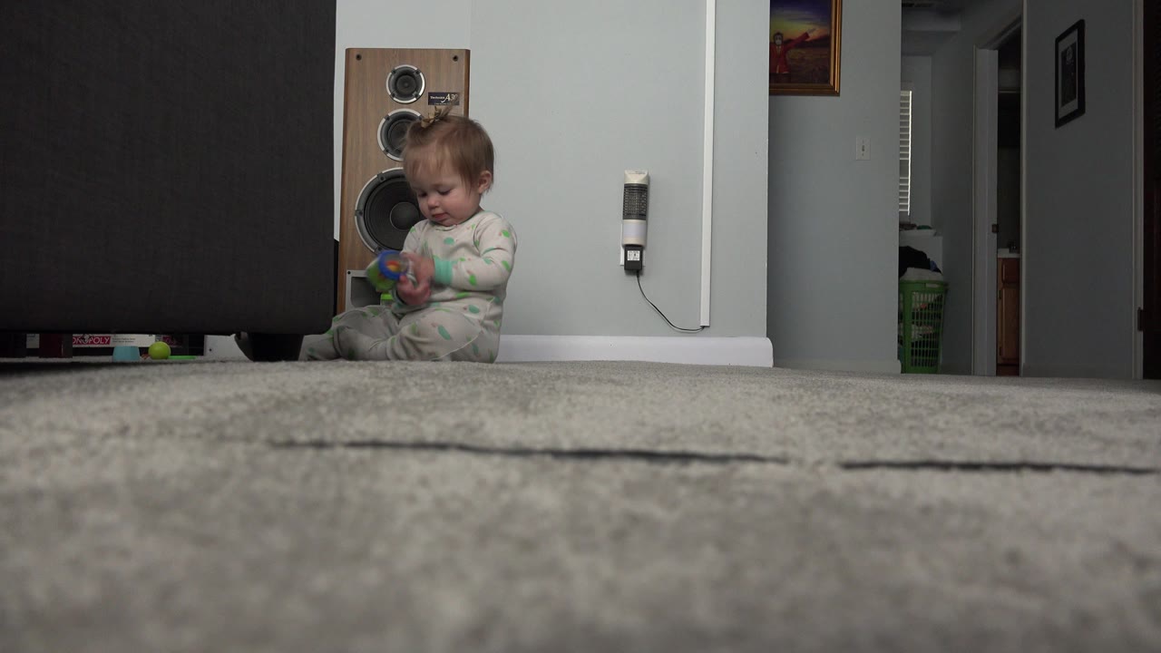 One second in, the baby in the green and white sleeper looks down at a toy beside the large speaker in the Westminster, Colorado home interior.