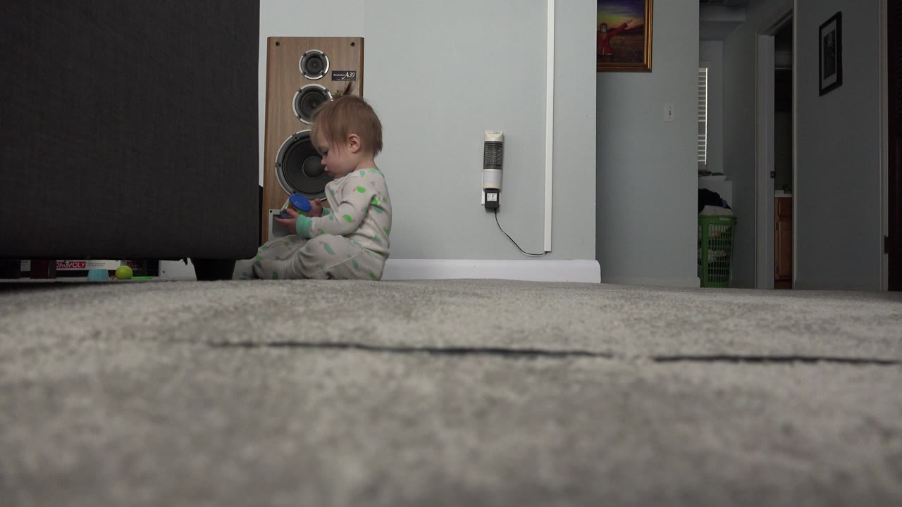 At 0:00 in Westminster, Colorado, a blonde baby girl sits in profile on the living room carpet beside a dark sofa while holding a small colorful object.