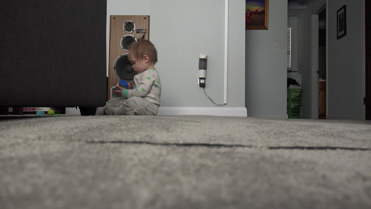 At 0:01 in Westminster, Colorado, the baby remains seated near the sofa, looking down as she handles the colorful object in the living room.