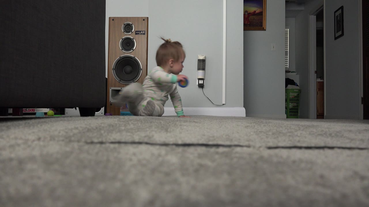 In a Westminster, Colorado home interior, a baby in patterned pajamas is low on a gray carpet beside a large speaker and dark furniture.