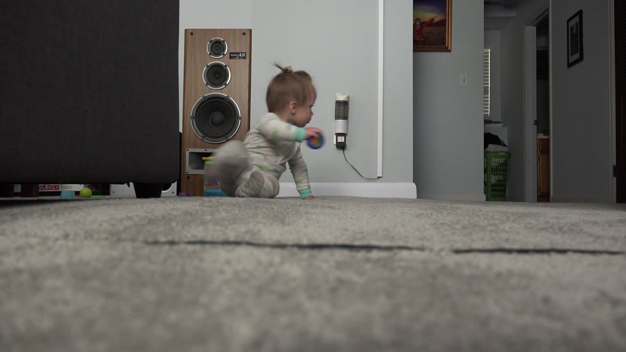 The baby crawls across the gray carpet in the Westminster home, with a wall-mounted device, speaker, hallway, and framed artwork behind them.