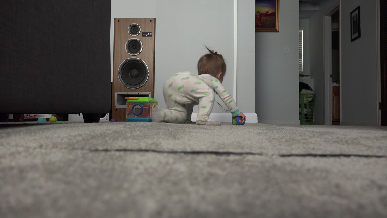 The infant leans forward on the carpet near a colorful toy, with the speaker, dark furniture, white baseboard, and hallway visible in the Westminster home.
