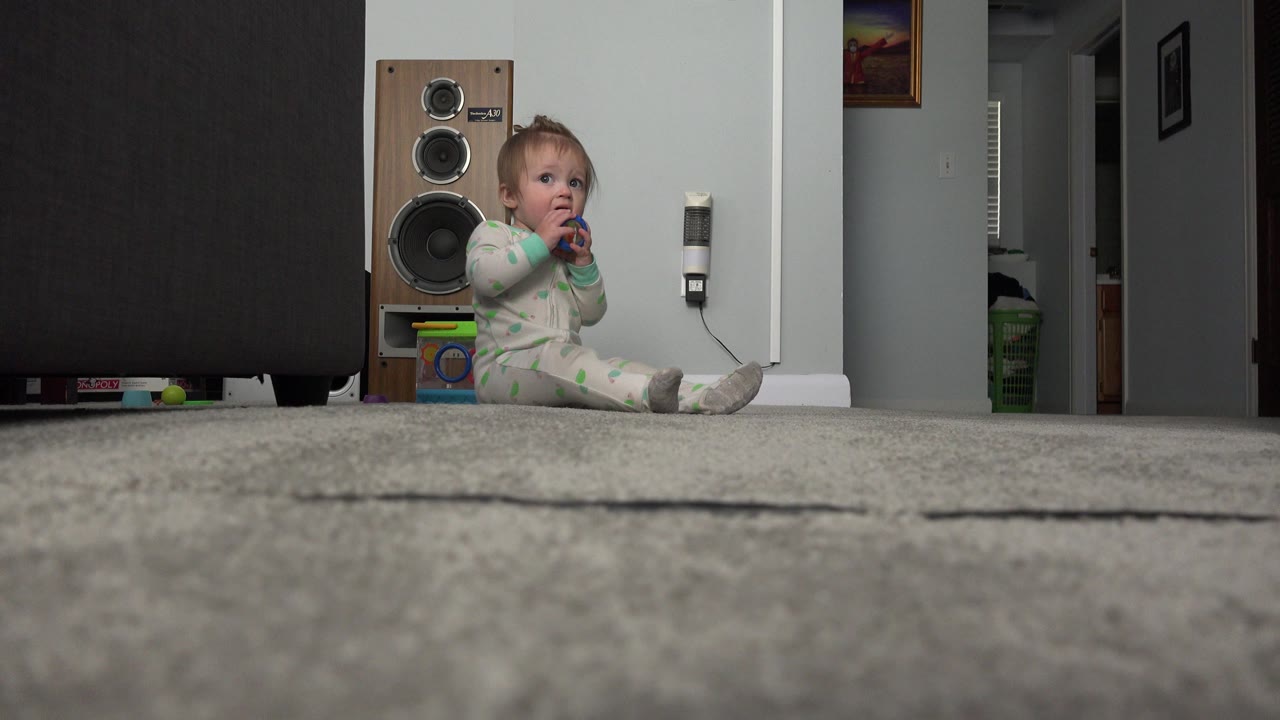 Baby sitting on carpet and biting a blue plastic toy near a large speaker.