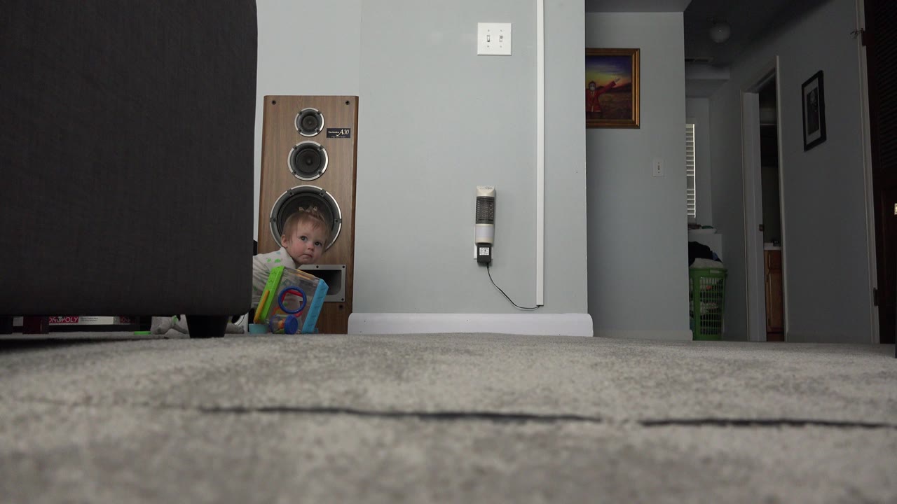 Baby peeking beside a wooden speaker while holding a toy on carpet.