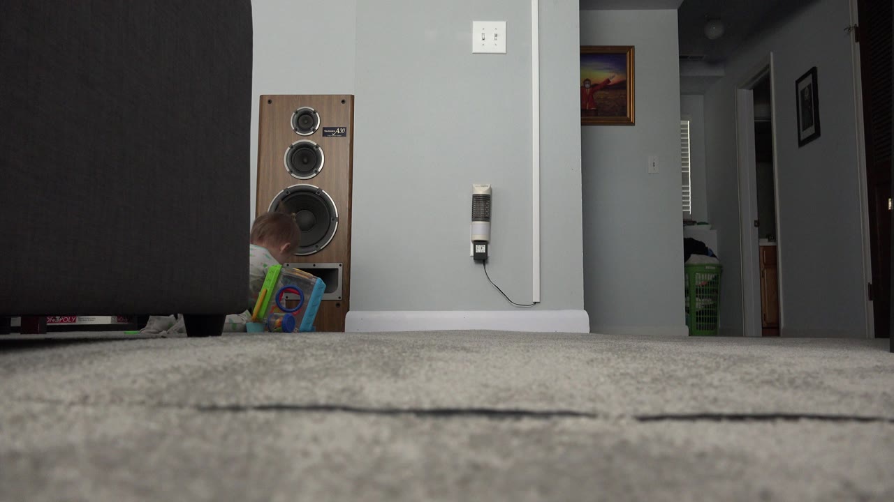 At 0:01, the baby remains low near the speaker and a colorful bag or toy, while the hallway and open dark doorway frame the background of the Westminster home interior.