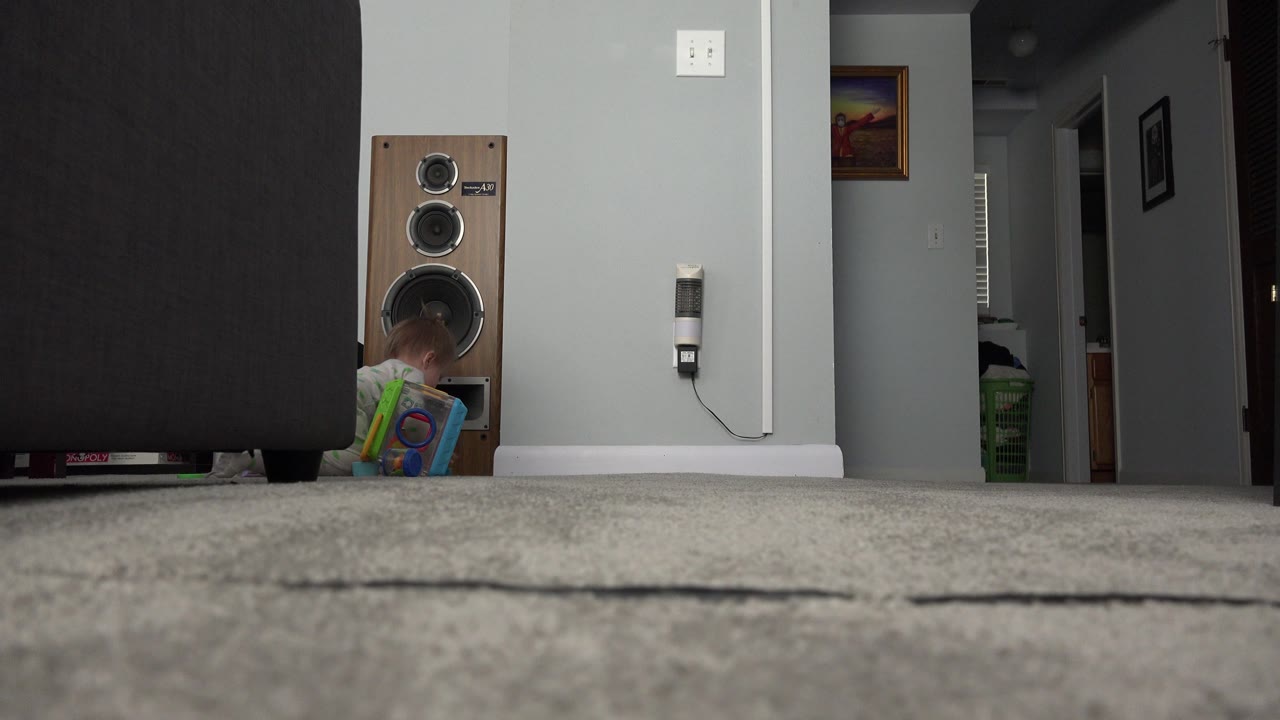 Baby partially hidden by a colorful object beside a large speaker on carpet.