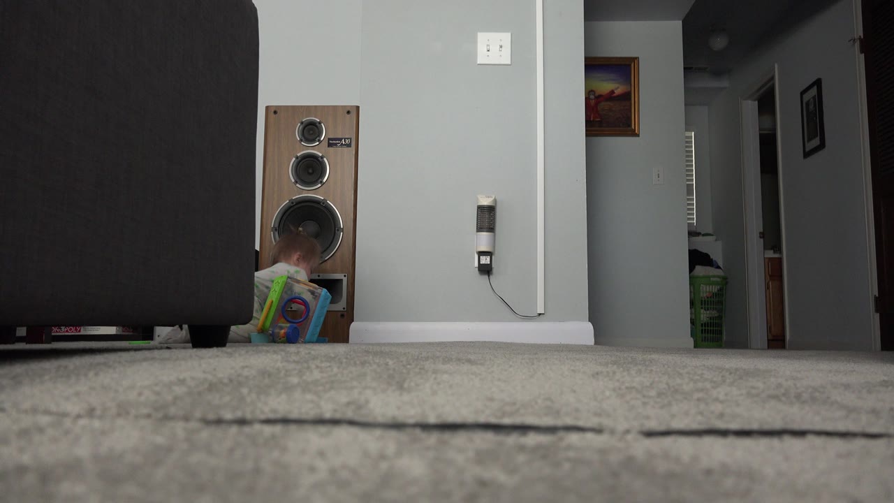 At 0:01, the child remains low and mostly hidden beside the speaker, with the dark sofa dominating the left foreground in the Westminster home interior.