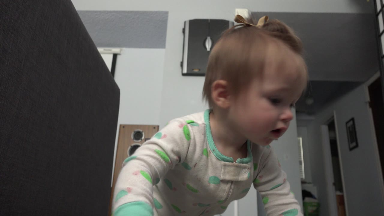 A blonde baby in white printed pajamas crawls across a Westminster, Colorado room, with dark furniture and a wall-mounted fixture behind her.