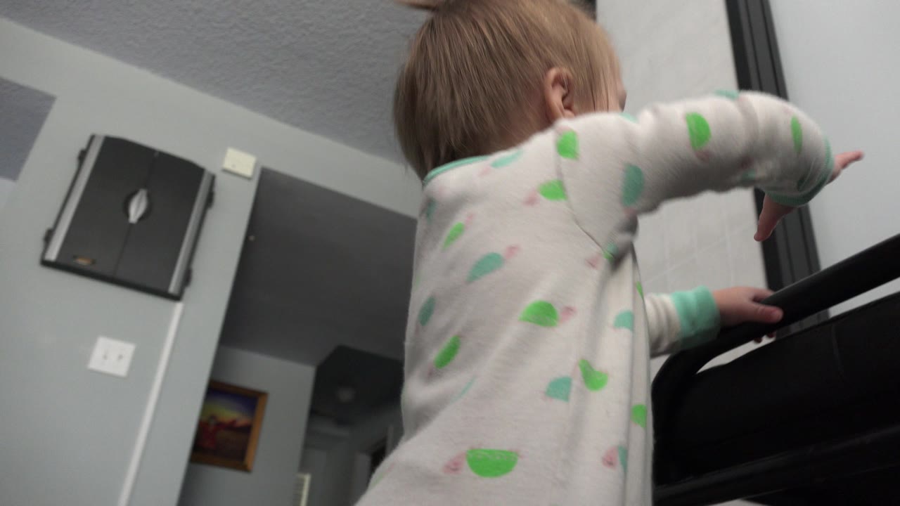 Toddler reaches over a black curved object indoors.