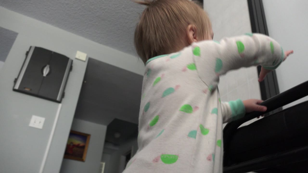 At 0:01 in Westminster, Colorado, the child keeps both hands near the top of the black object as the low tilted view shows gray walls and a doorway behind her.