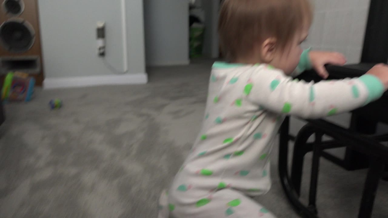 Baby in patterned pajamas holding a black chair in a carpeted room.