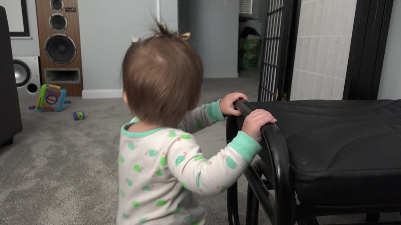 The infant remains at the black chair, seen from behind at floor level with speakers and small toys visible across the gray carpet.