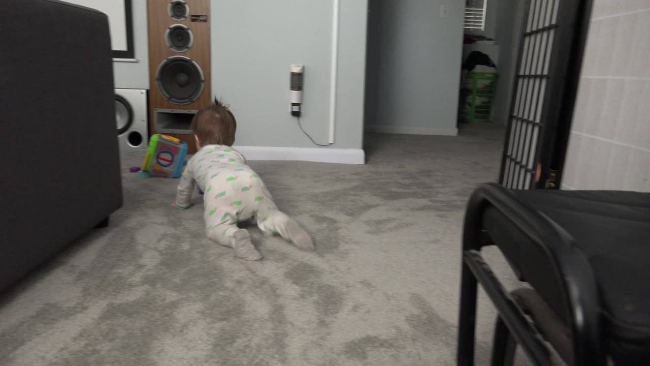 Baby crawling on gray carpet beside a dark bed or sofa.