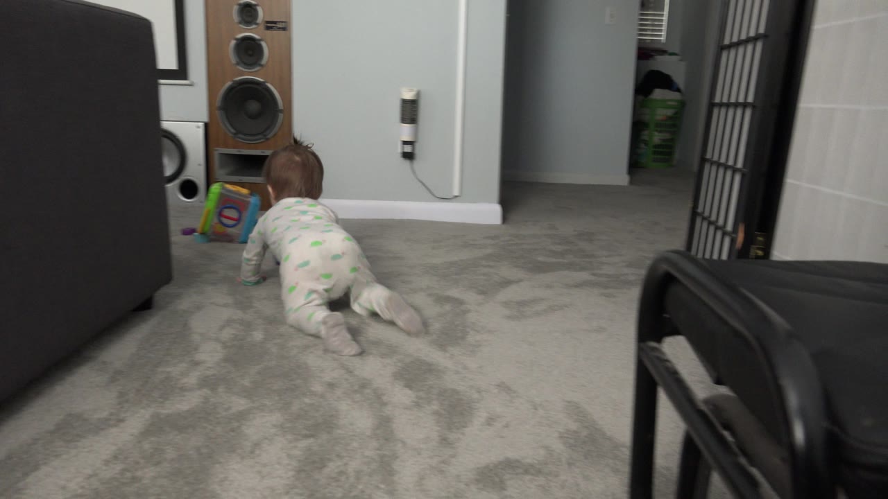 The baby continues crawling across the gray carpet in the Westminster, Colorado interior, moving closer to the tall wooden speaker and toy near the wall.