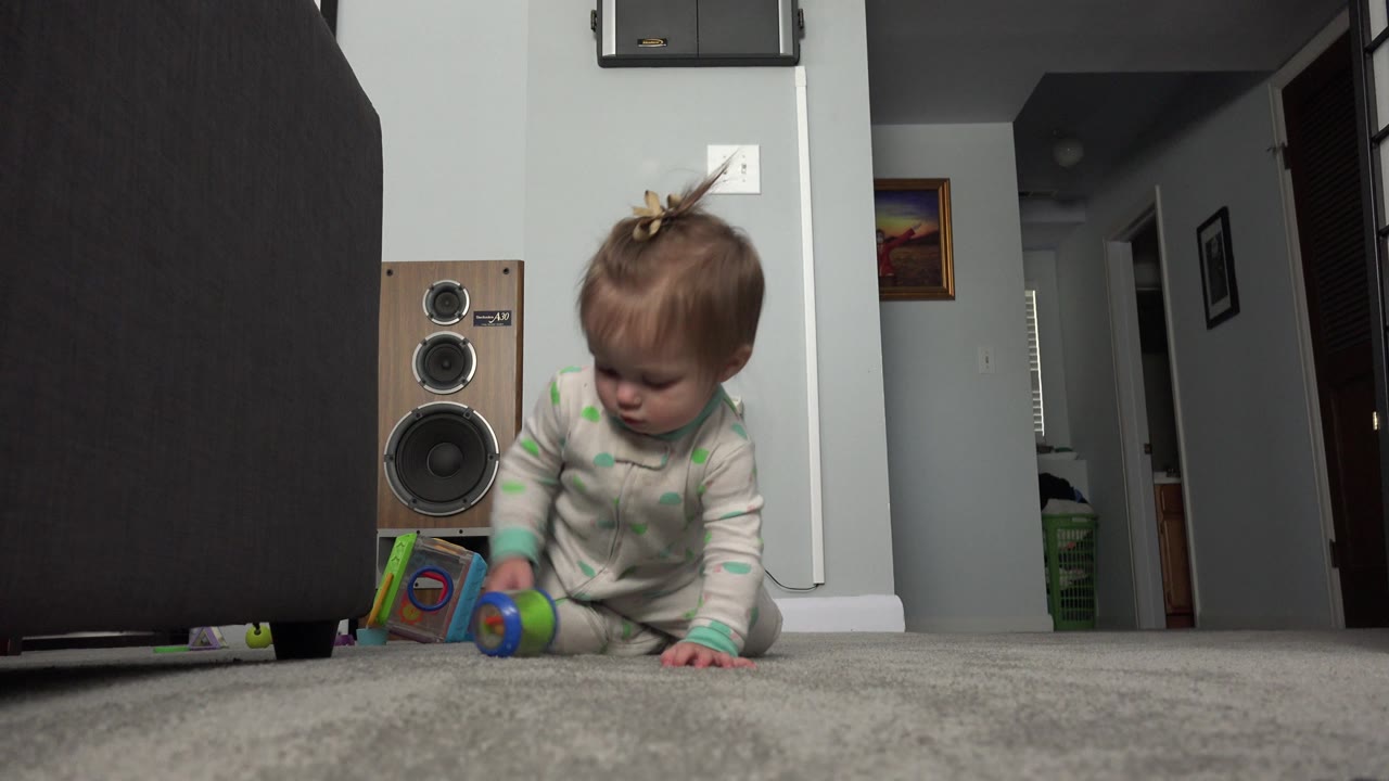 The baby leans forward on one hand near a small rolling toy on the carpet, with the floor-level camera showing the room wall, speaker, and hallway in Westminster, Colorado.