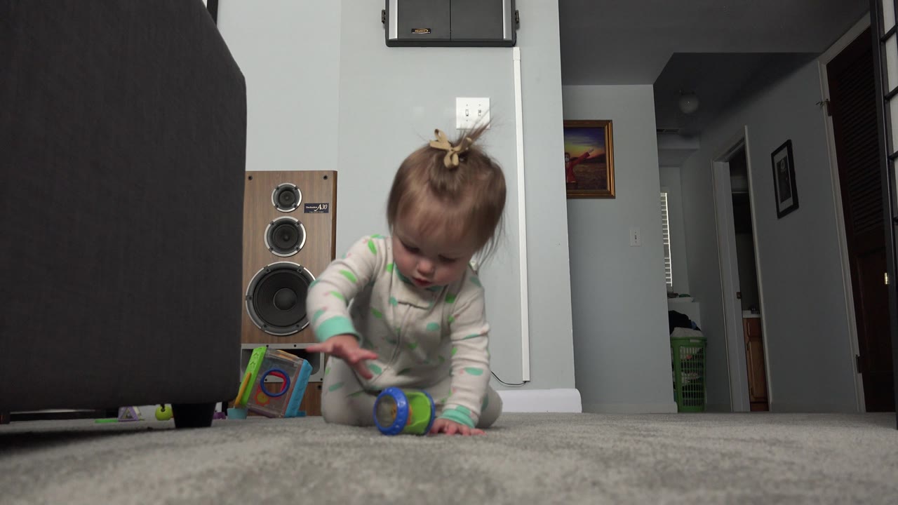 A baby kneels and reaches down to a colorful toy on the carpet, framed low against a pale wall, wood speaker, dark furniture, and hallway inside a Westminster, Colorado home.