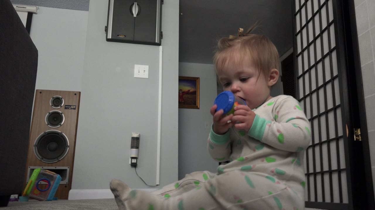 The baby remains seated on the carpet in the Westminster, Colorado interior, looking down at a blue toy while the tall speaker and pale wall stay visible behind them.