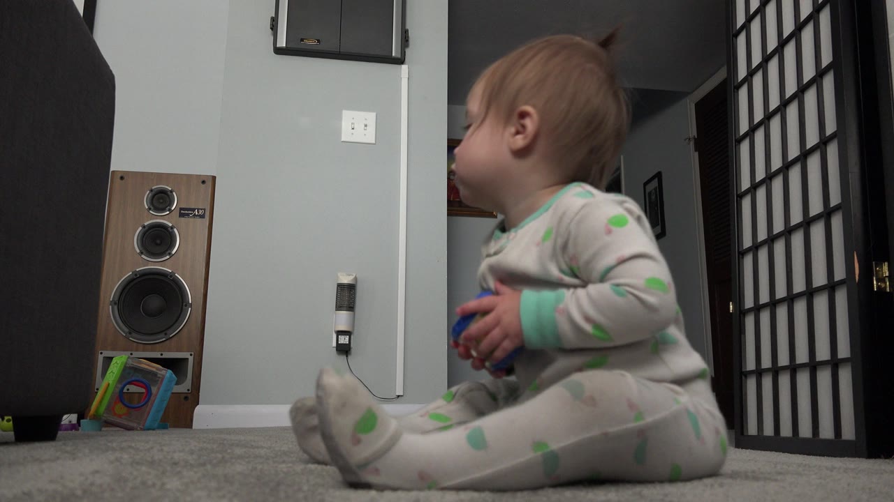 A blonde baby in dotted pajamas sits on gray carpet in a Westminster, Colorado room, holding a small colorful toy near a speaker and black room divider.