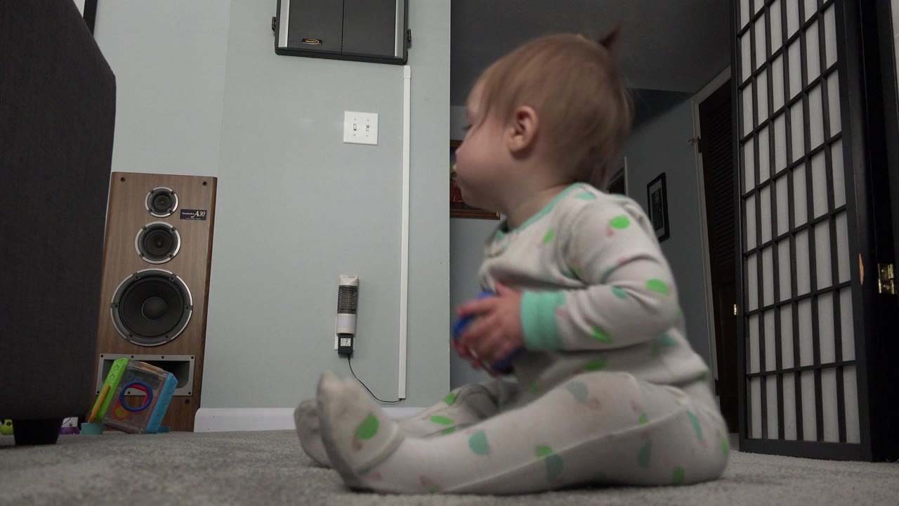 The baby sits in side profile on carpet in the Westminster, Colorado interior, with a wood speaker, wall switch, doorway, and black folding divider visible behind them.