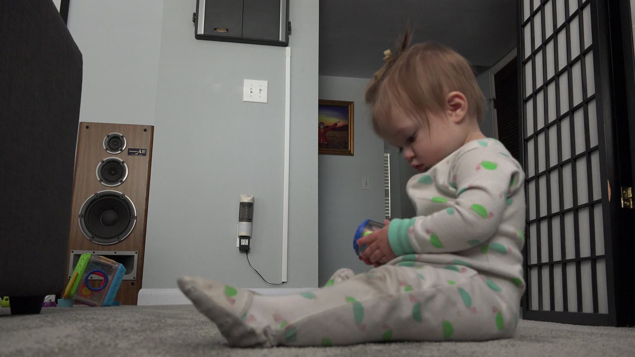 The child remains seated on the carpet in the Westminster home, holding the toy near their lap while the speaker, outlet, books, divider, and doorway stay visible behind them.