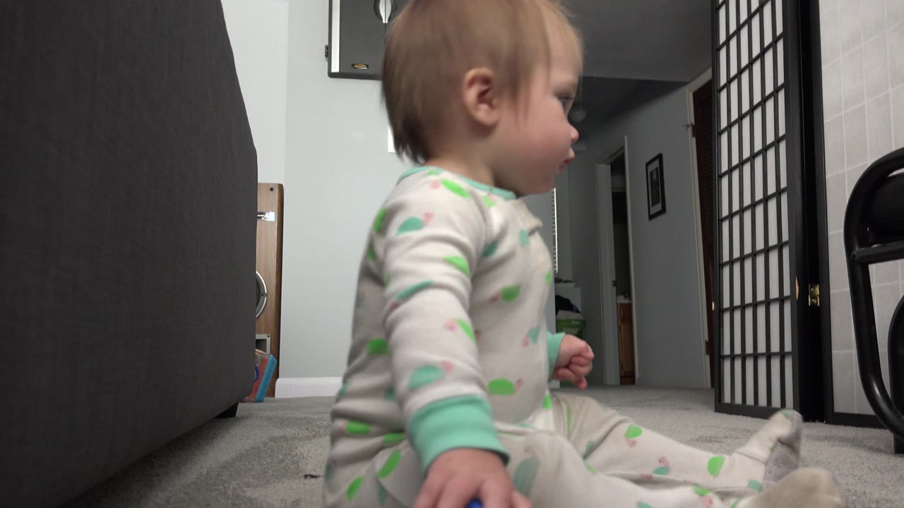 The baby remains seated in profile on the carpet, framed between dark furniture in the foreground and an open doorway deeper inside the Westminster home.