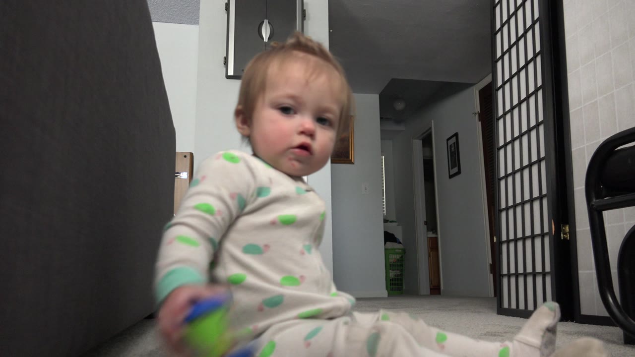 Toddler sitting on carpet near dark furniture and holding a colorful toy.