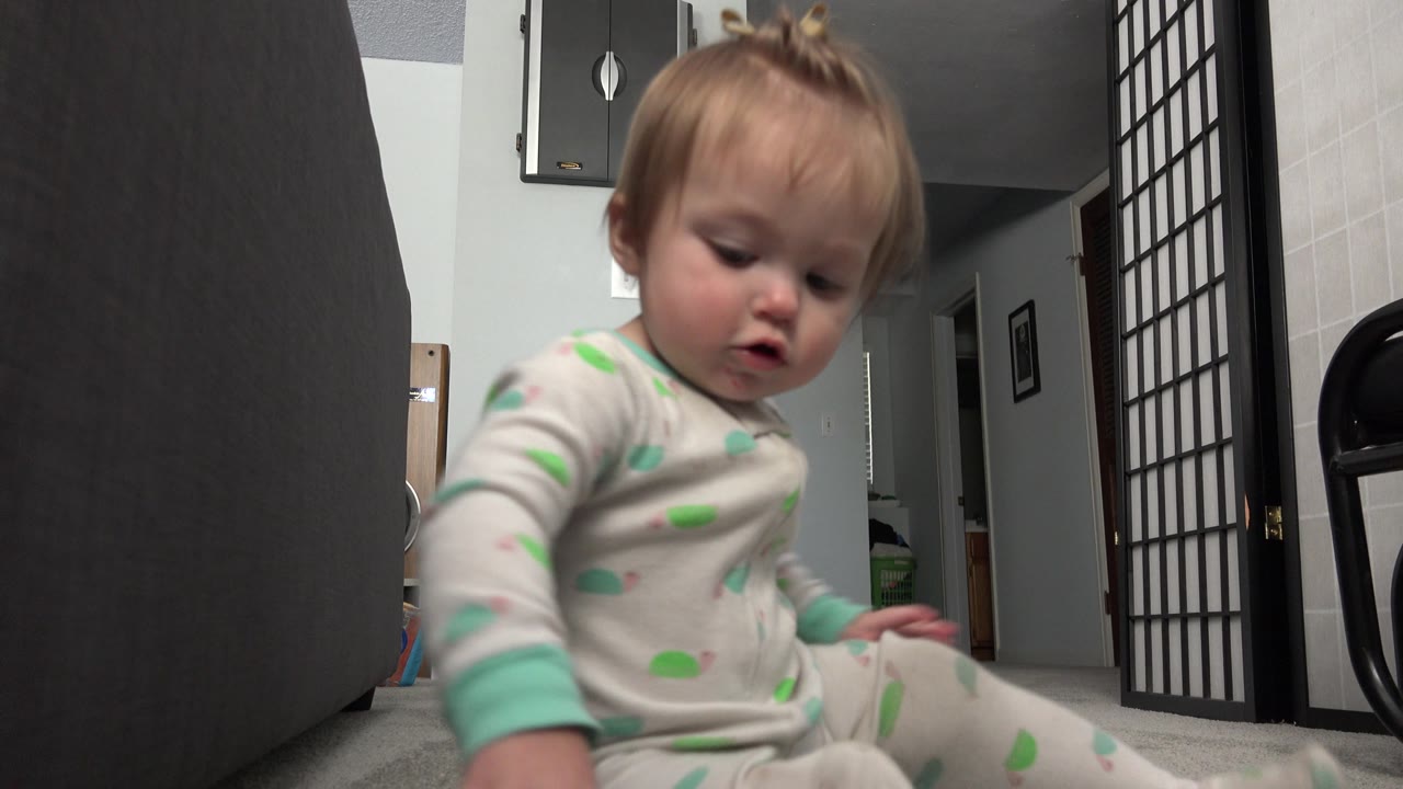 A seated toddler in light pajamas looks down on the carpet in a Westminster, Colorado interior, with a folding screen and chair frame visible behind.